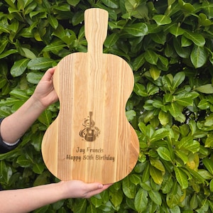 May include: A light brown wooden serving board shaped like a guitar. The board features a laser-engraved graphic of a guitar surrounded by swirling lines, with the text "Jay Francis" and "Happy 50th Birthday" engraved below the graphic.