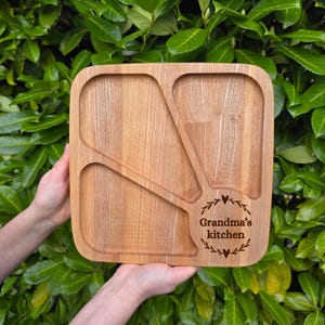 May include: A square wooden serving tray with three compartments and a decorative design. The tray is light brown with the text "Grandma's kitchen" in a circular design. The tray is held by two hands against a background of green leaves.