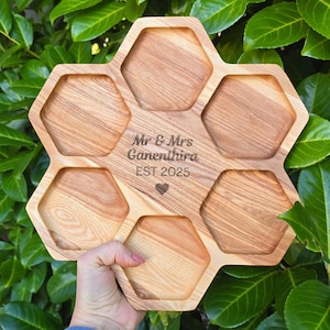 May include: A wooden serving tray with six hexagonal compartments. The tray is engraved with "Mr & Mrs Canenthira EST 2025" and a heart symbol. The tray is held against a backdrop of green foliage.