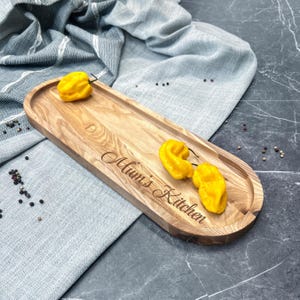 Puede incluir: Una bandeja de servir de madera larga y ovalada con un acabado de madera natural. La bandeja tiene bordes redondeados y está grabada con las palabras "Mum's Kitchen". Hay tres pimientos amarillos en la bandeja.