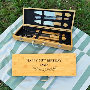 May include: A wooden barbecue tool set with stainless steel blades and light brown wooden handles. The set includes knives, a fork, and tongs, all stored in a bamboo case. The case lid reads "HAPPY 50TH BIRTHDAY DAD" with a decorative leaf design.