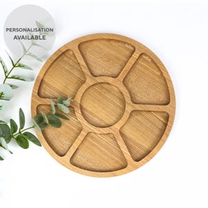 May include: A round, light brown wooden serving tray with six wedge-shaped compartments and a central circular area. The tray has a natural wood grain pattern and the text "PERSONALISATION AVAILABLE" is in a white circle.