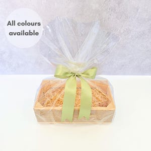 Può includere: Un cesto regalo in legno riempito di paglia, avvolto in cellophane trasparente e legato con un nastro di raso verde chiaro. Il testo "All colours available" è in un cerchio bianco.