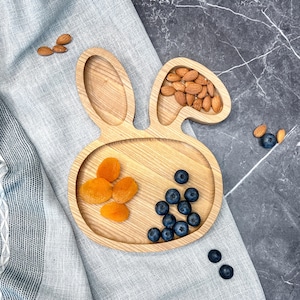 May include: A wooden bunny-shaped plate with three sections. The plate is filled with dried apricots, blueberries, and almonds.