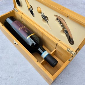 Peut inclure: Un coffret cadeau de vin en bambou avec une bouteille de vin rouge et des accessoires. L'ensemble comprend un tire-bouchon, un bouchon et un verseur. La boîte est ouverte, révélant le contenu.