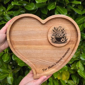 Può includere: Un vassoio di legno a forma di cuore con un sottobicchiere. Il vassoio è marrone chiaro con venature naturali del legno. Il sottobicchiere presenta un disegno floreale in bianco e nero attorno a una tazza da tè. Il testo "For Mum's sweet moments" è inciso sul vassoio.