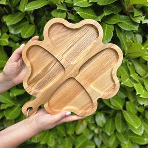 Op de afbeelding: Een houten serveerschaal in de vorm van een klavertjevier. De schaal is gemaakt van lichtgekleurd hout en heeft vier aparte compartimenten. Het heeft een handvat met een gat om op te hangen. De achtergrond is een weelderige groene struik.