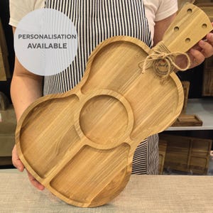 Può includere: Un vassoio da portata in legno a forma di chitarra con quattro scomparti separati. Il vassoio è realizzato in legno marrone chiaro e ha una finitura naturale. Il vassoio è tenuto da una mano. Il testo "PERSONALISATION AVAILABLE" è visibile nell'immagine.