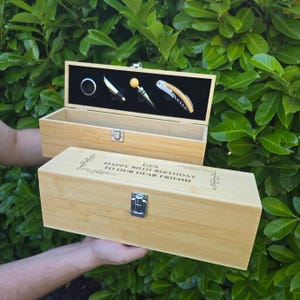May include: A light-coloured bamboo wine box with a metal latch, engraved with the text GUS HAPPY 80TH BIRTHDAY TO OUR DEAR FRIEND. The open lid reveals a black interior holding a silver ring, a small knife, a bottle stopper, and a corkscrew.