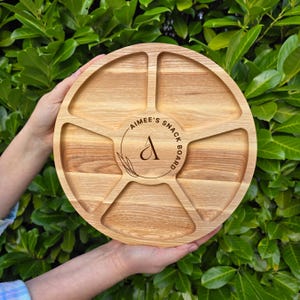 May include: A round, light-colored wooden snack board with six compartments. The center of the board features the text "Aimee's Snack Board" in a circular design with a stylized "A". The board is held by two hands against a backdrop of green foliage.