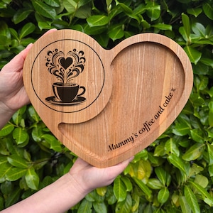 Può includere: Vassoio di legno a forma di cuore con un inserto circolare con un disegno di tazza di caffè. Il vassoio è inciso con "Mummy's coffee and cake x". Il legno ha una venatura naturale e un colore marrone chiaro.