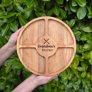 May include: A round, wooden serving tray with four compartments and a central circular area. The tray is engraved with the text "Grandma's kitchen" and crossed utensils. The tray is held by two hands against a backdrop of green foliage.