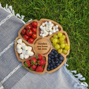 May include: A wooden serving platter with six compartments, filled with fresh food items. The platter contains cherry tomatoes, mozzarella balls, strawberries, feta cheese, green grapes, and blueberries. The center of the platter has the text "Camilla's new home est. 2026".