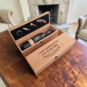 May include: A rectangular light wood wine box with a hinged lid, engraved with the text CHRIS ROBERTSON EST. 18/01/1986 and a leaf design. The open box contains a wine bottle and four metal wine accessories set in a black foam insert.