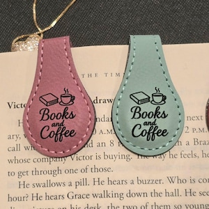 May include: Two leather bookmarks, one pink and one teal, with the words "Books and Coffee" printed on them. Each bookmark has a stitched border and a design of a book and a coffee cup.