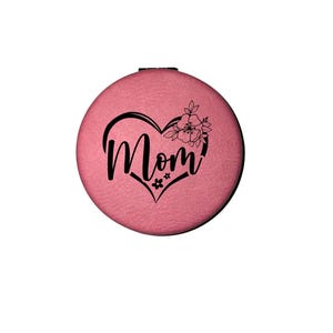May include: A round, pink compact mirror with a black heart-shaped design that includes the word "Mom" in cursive script. The heart is adorned with floral accents, creating a personalized and decorative accessory.