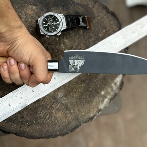 May include: A black chef's knife with a wooden handle is held next to a metal ruler. The knife blade has a logo etched on it. A silver watch with a black band is in the background.