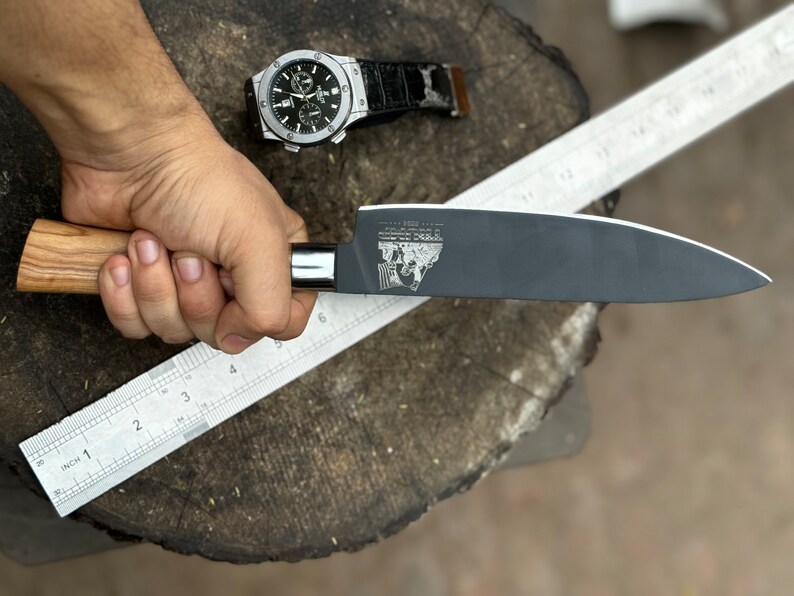 May include: A black chef's knife with a wooden handle is held next to a metal ruler. The knife blade has a logo etched on it. A silver watch with a black band is in the background.