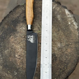 May include: A black chef's knife with a wooden handle, next to a ruler. The blade is approximately 10 inches long. The handle is light brown with a wood grain pattern. The knife is on a wooden surface.