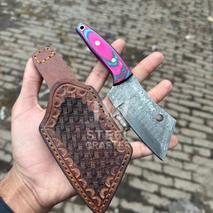 May include: A small cleaver knife with a pink and blue patterned handle and a patterned blade. The knife is held next to a brown leather sheath with a woven design. The text "Blue Angel Forge" is visible on the blade.
