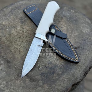 May include: A handcrafted knife with a white handle and a patterned blade, in a black leather sheath with yellow stitching. The knife is on a wooden surface, with the text "STEEL RAFTERS" visible.