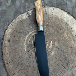 May include: A chef's knife with a black blade and a light-colored wooden handle. The handle has a natural, textured appearance. A silver metal bolster separates the blade from the handle. The knife is resting on a wooden surface.