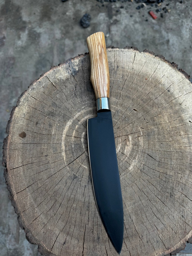 May include: A chef's knife with a black blade and a light-colored wooden handle. The handle has a natural, textured appearance. A silver metal bolster separates the blade from the handle. The knife is resting on a wooden surface.