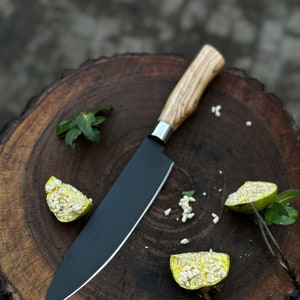 May include: A chef's knife with a black blade and a light wood handle, resting on a dark wood cutting board. The knife is next to sliced citrus fruit. The handle is made of light-colored wood.