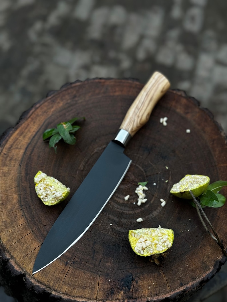 May include: A chef's knife with a black blade and a light wood handle, resting on a dark wood cutting board. The knife is next to sliced citrus fruit. The handle is made of light-colored wood.