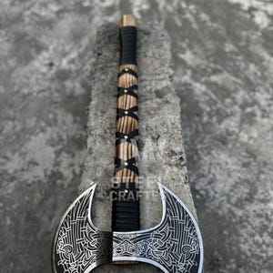 May include: A double-bladed axe with a wooden handle wrapped in black leather and adorned with brass accents. The silver blades feature intricate etched designs. The text "STEEL CRAFTE" is visible on the handle.