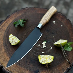 May include: A black chef's knife with a wooden handle rests on a dark brown wooden cutting board. The knife has a silver bolster and a sharp blade. Sliced fruit and green leaves are scattered around the knife.