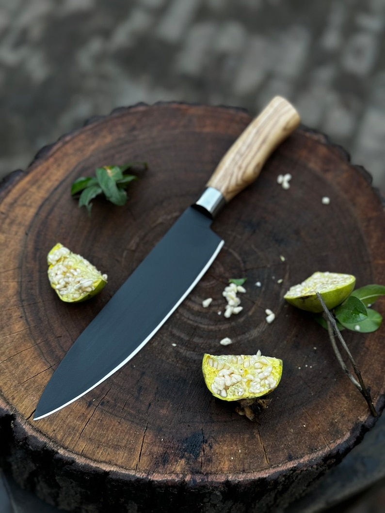 May include: A black chef's knife with a wooden handle rests on a dark brown wooden cutting board. The knife has a silver bolster and a sharp blade. Sliced fruit and green leaves are scattered around the knife.