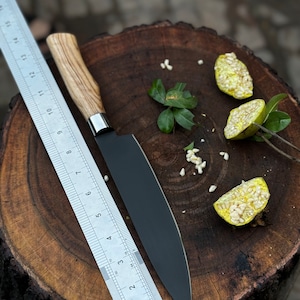 May include: A black chef's knife with a wooden handle and a stainless steel bolster rests on a wooden surface next to a metal ruler. The knife blade is approximately 8 inches long. Sliced fruit and leaves are also present.