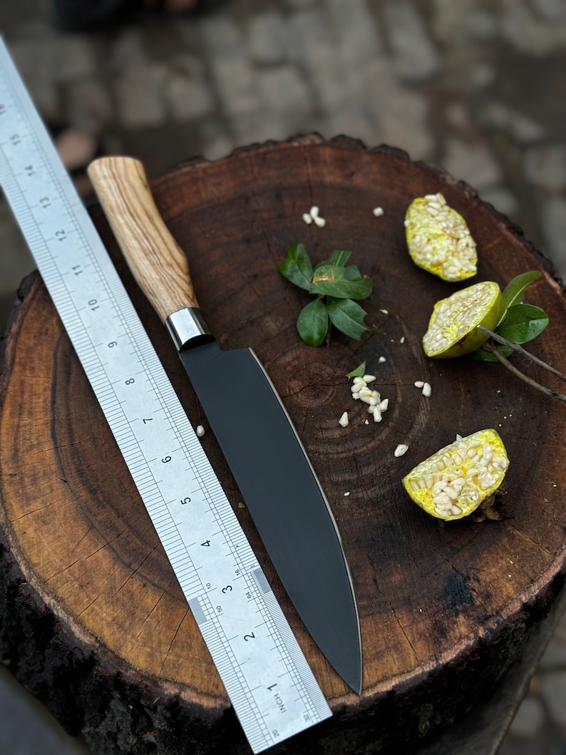 May include: A black chef's knife with a wooden handle and a stainless steel bolster rests on a wooden surface next to a metal ruler. The knife blade is approximately 8 inches long. Sliced fruit and leaves are also present.