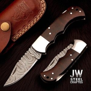 May include: Two folding knives with dark wood handles and patterned steel blades are displayed. One knife is open, revealing the blade, while the other is closed. A brown leather sheath with a button closure is visible in the background. The text "JW STEEL CRAFTES" is also visible.