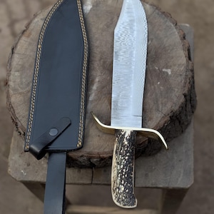 Könnte beinhalten: Ein großes, verziertes Messer mit gemusterter Klinge und einem Griff aus Geweihmaterial. Das Messer hat einen Messing-Handschutz und wird von einer schwarzen Lederscheide mit beigen Nähten begleitet. Das Messer und die Scheide werden auf einer Holzoberfläche präsentiert.