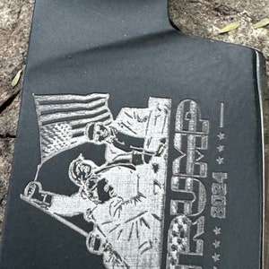May include: A black metal cleaver with an engraved design. The design features an American flag, figures, and the word "TRUMP" with the year "2024" and stars.