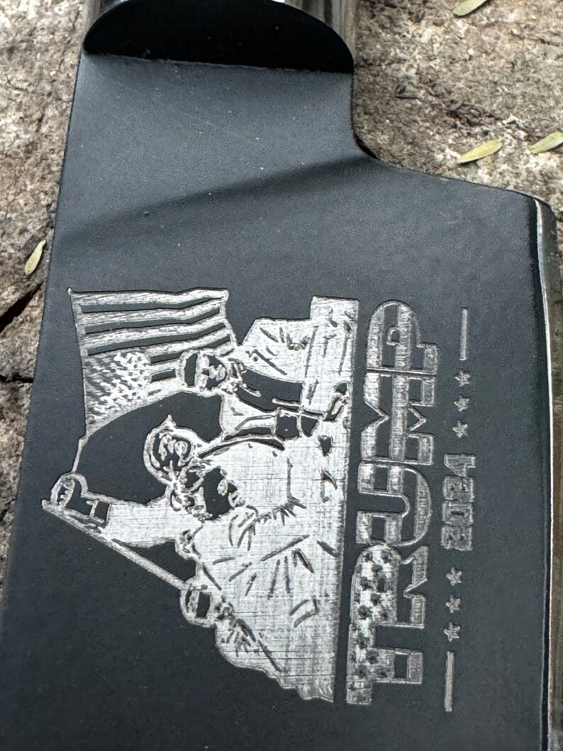 May include: A black metal cleaver with an engraved design. The design features an American flag, figures, and the word "TRUMP" with the year "2024" and stars.