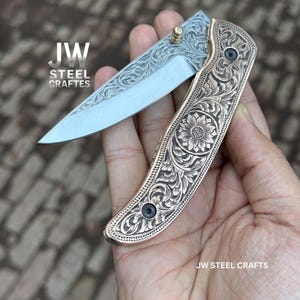 May include: A partially open folding knife with a silver blade and a bronze handle. The handle features intricate floral engravings. The text "JW STEEL CRAFTES" is visible.