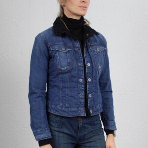 May include: A woman wearing a blue denim jacket with a black faux fur collar, a black turtleneck shirt, blue jeans, and white trainers.