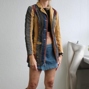May include: A patchwork blazer with vertical panels of denim, brown, and tan fabric. The blazer has a notched collar and button closure. The outfit includes a denim mini skirt and knee-high suede boots. The background is a plain white wall.