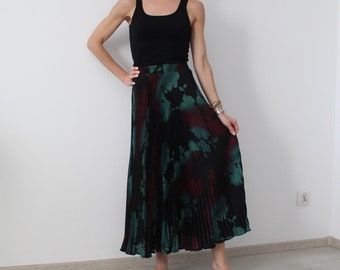 May include: A woman wearing a black tank top and a long, pleated skirt with a green and black floral print. The skirt has a high waist and falls to her ankles.