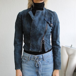 May include: A dark denim jacket with an off-centre zip and a small pocket. The jacket is worn with light wash denim jeans. The model is wearing a black roll neck.