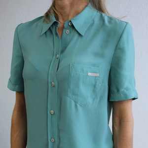 Pode incluir: Uma camisa azul turquesa de manga curta com botões e um pequeno bolso no peito esquerdo. A camisa tem um fecho de botões prateados e uma etiqueta prateada com a palavra "GINA" no bolso.