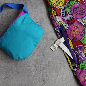 May include: A turquoise and purple nylon fanny pack with a blue adjustable strap. The fanny pack is lying on a gray textured surface.  The fanny pack is next to a colorful fabric with a pattern of license plates.