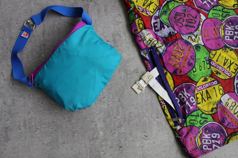 May include: A turquoise and purple nylon fanny pack with a blue adjustable strap. The fanny pack is lying on a gray textured surface.  The fanny pack is next to a colorful fabric with a pattern of license plates.