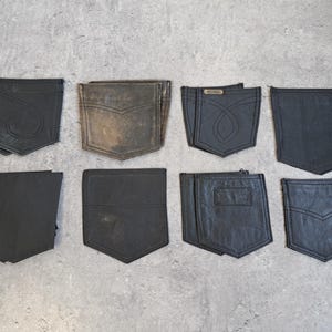 May include: An assortment of eight black leather pockets, likely from jeans or other garments, are arranged on a gray surface. The pockets vary in style, with some featuring decorative stitching and unique shapes.
