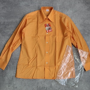 Könnte beinhalten: Ein orangefarbenes langärmliges Button-Down-Hemd mit spitzem Kragen. Das Hemd hat ein Etikett, das am oberen Knopf befestigt ist. Das Hemd ist teilweise von einer durchsichtigen Plastikfolie bedeckt. Das Hemd liegt auf einer grauen Oberfläche.