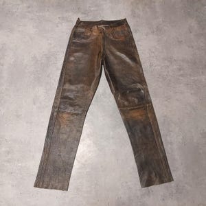 May include: A pair of dark brown leather trousers featuring a distressed, weathered finish with visible grain texture. The trousers include a standard waistband with a button closure, two front pockets, and straight-cut legs with vertical seam detailing.
