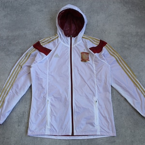 May include: White windbreaker jacket with gold and red accents. The jacket has a zipper closure and a hood. The jacket has the Adidas logo and the Spanish national team crest.